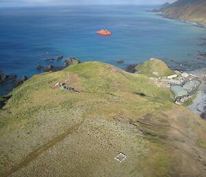 Aerial view of some small shipping containers and apple hut on top of a hill overlooking Macquarie Island research station, with the ship Aurora Australis in the distant harbour.