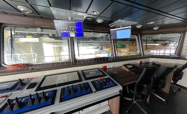 Inside of a room with control panels, desk, chairs and display screens, with large windows overlooking the science deck.