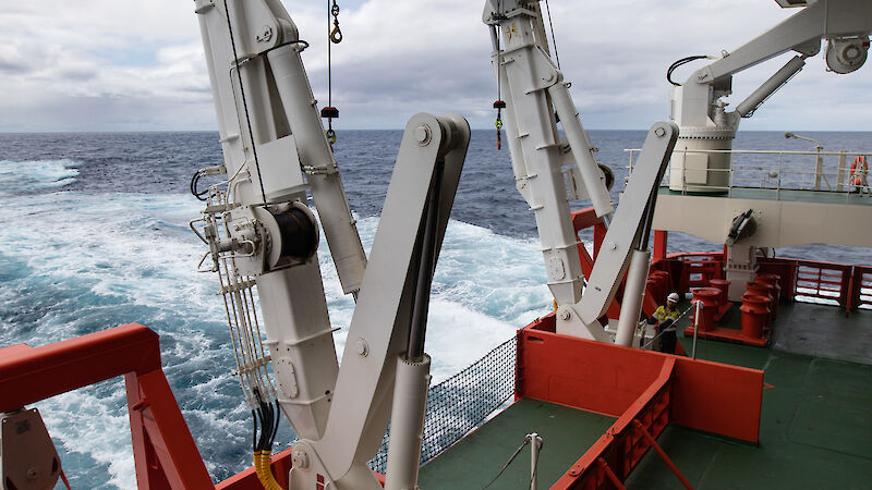 Orange and white machinery on the green deck of a ship. Behind the ship is a pale blue wake.