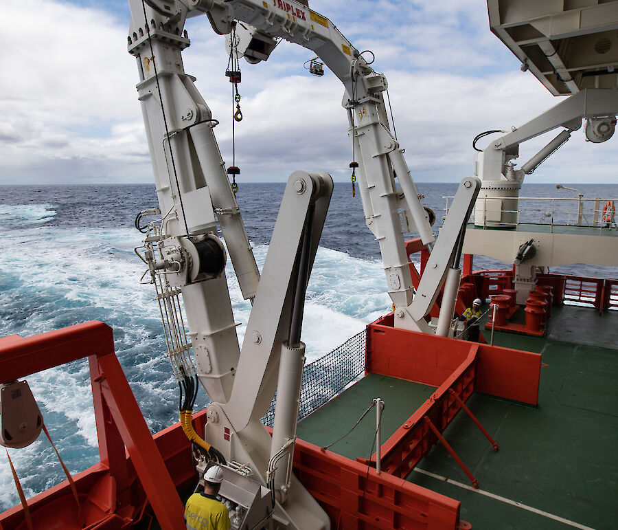 Orange and white machinery on the green deck of a ship. Behind the ship is a pale blue wake.