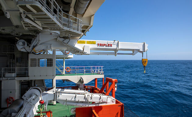 Machinery on the green deck of a ship. A large white crane is attached to the roof of the space.