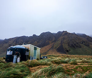 A field hut and tank on grass at the base of a mist covered hill