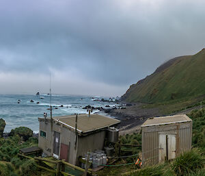 Two small huts at the base of a hill with the grey ocean in the background