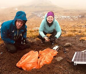 Two expeditioners squatting next to a solar panel in the field
