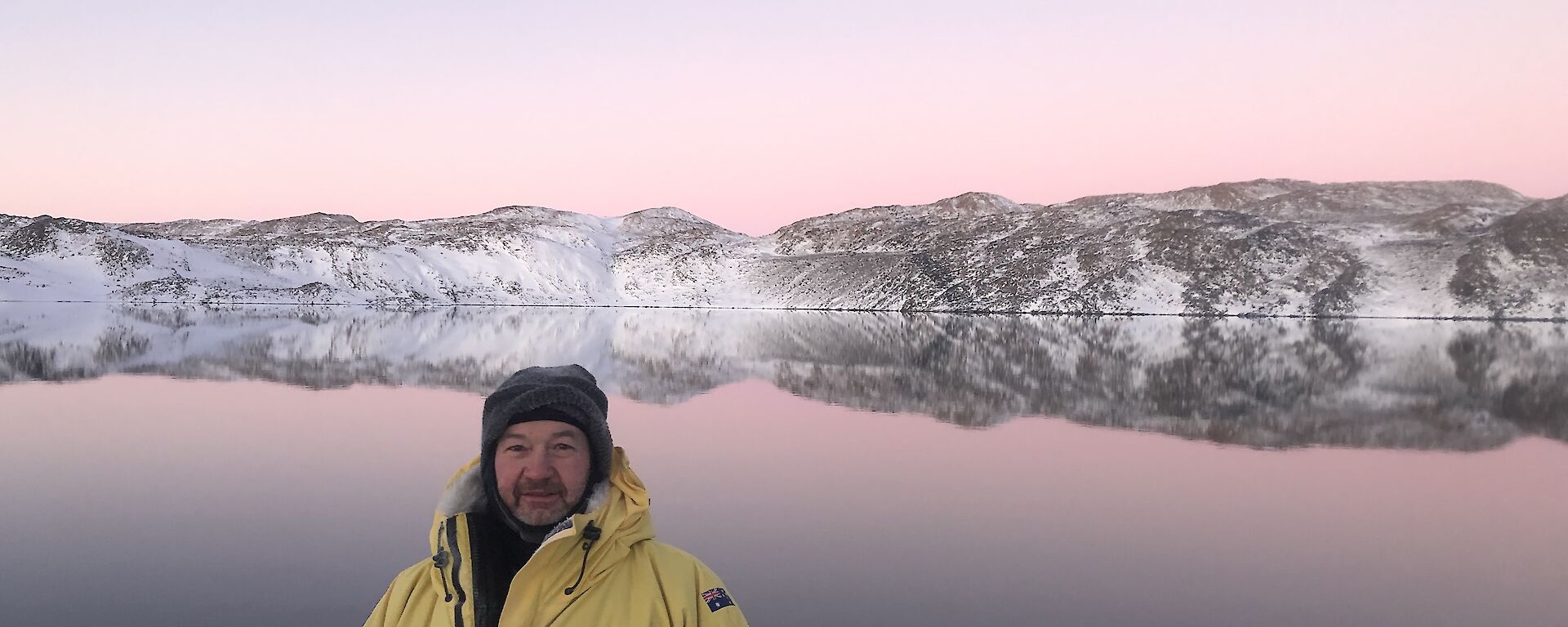 A man poses in front of Deep Lake.  The mountains behind are reflected perfectly in the water.