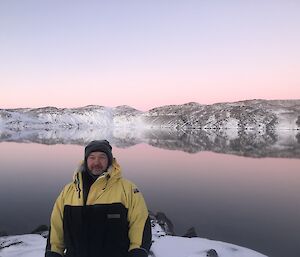 A man poses in front of Deep Lake.  The mountains behind are reflected perfectly in the water.