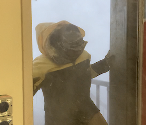A man walking in through a door.  Blizzard conditions can be seen outside and his hood is being blown up over his head.