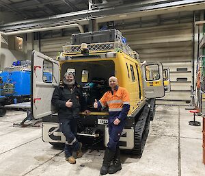 Two men stand at the back of a yellow Hagglund in a workshop giving a thumbs up
