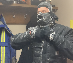 A man pulling down the zip on his jacket, which is covered in snow.  He is wearing a hat, gloves and scarf up over his face.