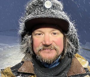 A selfie of a man in a woolly hat with icicles on his facial hair. Snow is falling in the background.