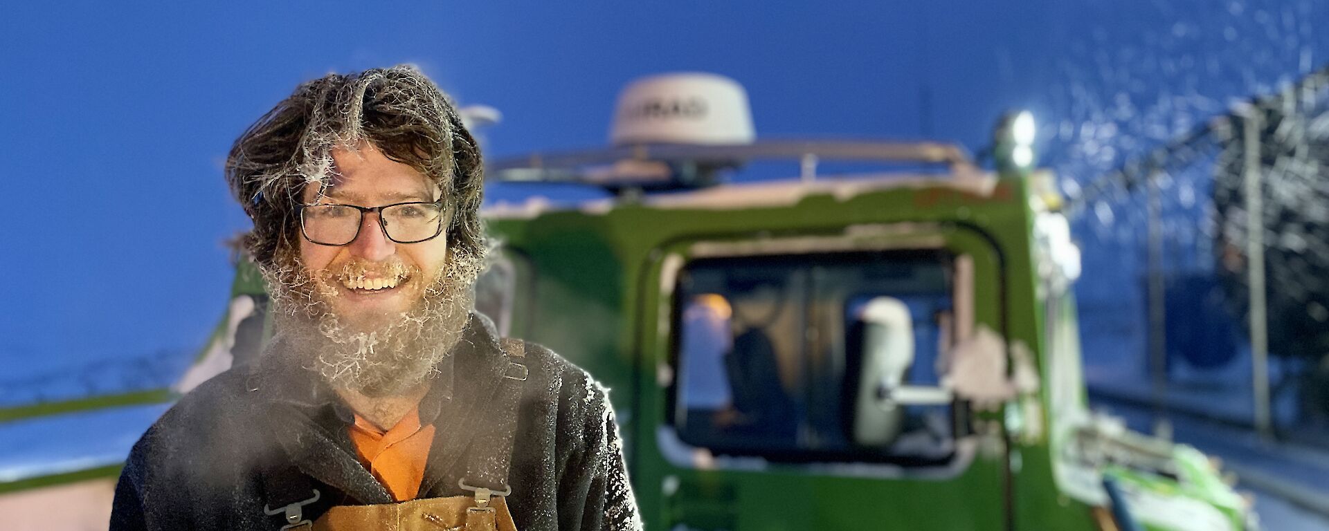 A man smiling to camera.  His beard and hair is covered in ice and his breath is clouding in the cold air.  A green Hagglund is behind him.