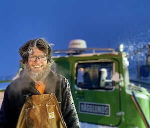 A man smiling to camera.  His beard and hair is covered in ice and his breath is clouding in the cold air.  A green Hagglund is behind him.