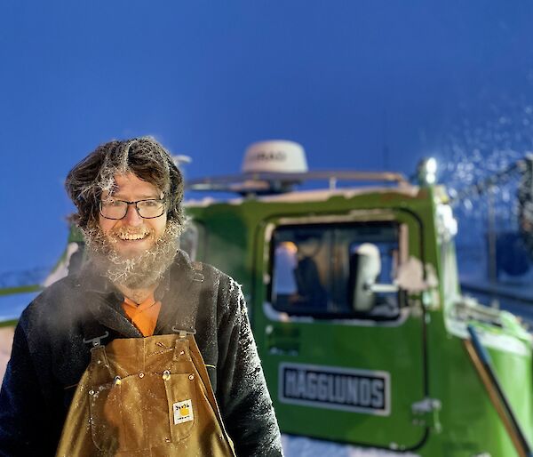 A man smiling to camera.  His beard and hair is covered in ice and his breath is clouding in the cold air.  A green Hagglund is behind him.