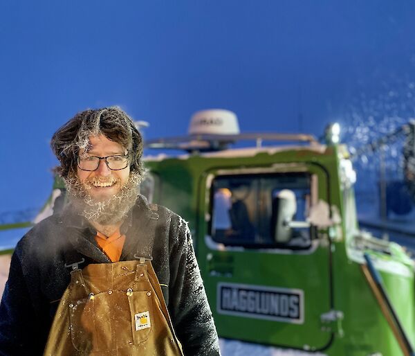 A man smiling to camera. His beard and hair is covered in ice and his breath is clouding in the cold air. A green Hagglund is behind him.