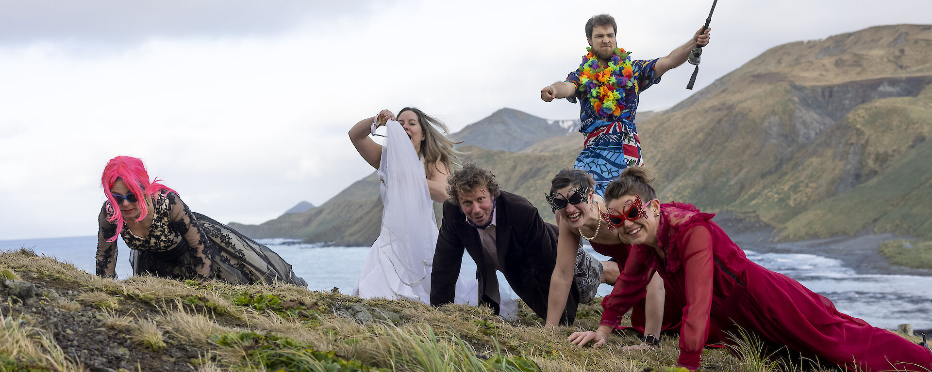 Five people in the tussoch grass doing push ups, dressed in evening gowns and suits.  A man behind, wearing a Hawaiian shirt holds a fishing rod aloft,