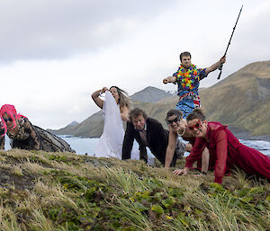 Five people in the tussoch grass doing push ups, dressed in evening gowns and suits.  A man behind, wearing a Hawaiian shirt holds a fishing rod aloft,