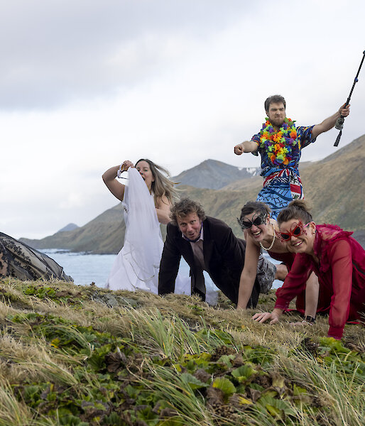 Five people in the tussoch grass doing push ups, dressed in evening gowns and suits.  A man behind, wearing a Hawaiian shirt holds a fishing rod aloft,
