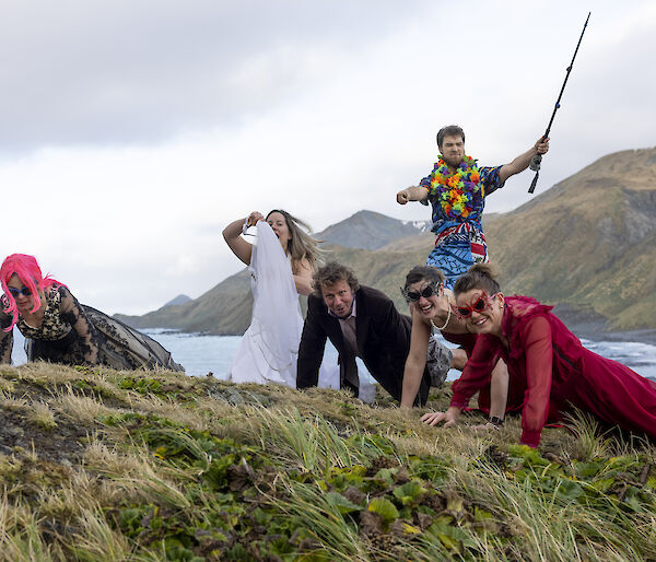 Five people in the tussoch grass doing push ups, dressed in evening gowns and suits.  A man behind, wearing a Hawaiian shirt holds a fishing rod aloft,