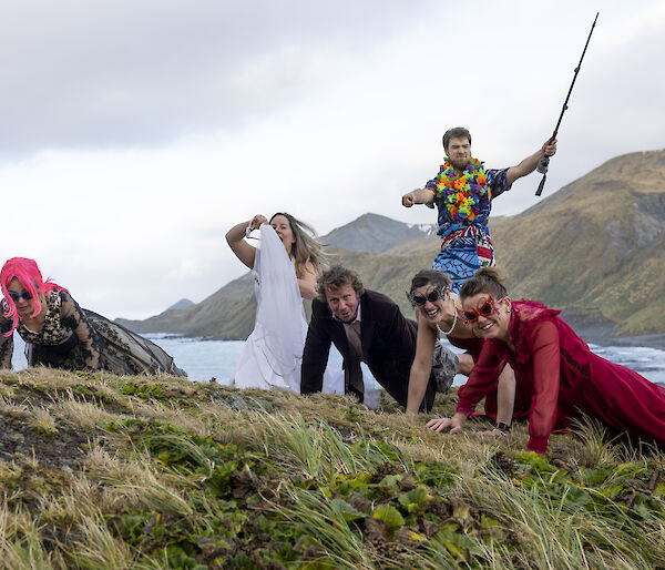 Five people in the tussoch grass doing push ups, dressed in evening gowns and suits. A man behind, wearing a Hawaiian shirt holds a fishing rod aloft,
