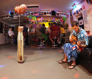 A man in Hawaiian shirt and sarong sits on a couch playing the guitar.  A large wooden leopard ornament is on the dance floor in front of him with balloons round its neck.  Other people are seen at the bar in the background.