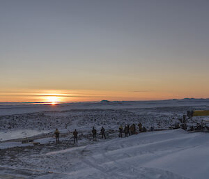 A view across a snowy landscape towards an orange sunrise.  A group of people can been seen watching the sunrise in the dusk.