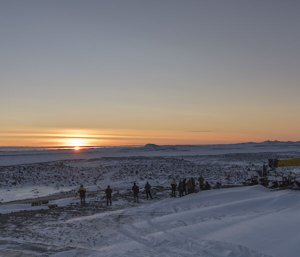 A view across a snowy landscape towards an orange sunrise.  A group of people can been seen watching the sunrise in the dusk.
