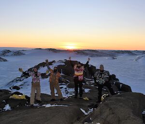 Four expeditioners in Hawaiian shirts, arms raised in the air smiling to camera with a snow covered landscape and sunrise behind them.