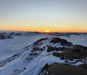 A snow covered landscape looking across some rocks, over a frozen lake, to a sunrise on the horizon