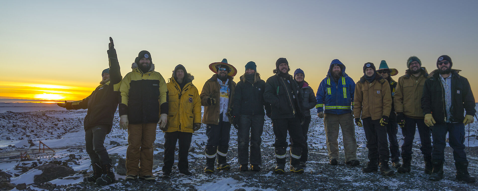 A portrait shot of a group of expeditioners, some in mexican hats, standing in a line smiling to camera with the sunrise behind them.