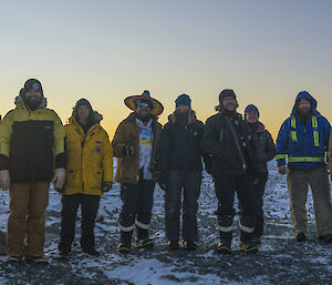 A portrait shot of a group of expeditioners, some in mexican hats, standing in a line smiling to camera with the sunrise behind them.