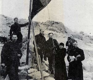An old black and white image of a group of  expeditioners raising a Norwegian flag on a wooden flagpole and looking to camera