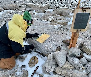 An expeditioner kneeling next to the Proclamation Point cairn about to write their name in the visitors book.