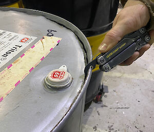 A man using the tin-opener feature on a multi-tool to prise the lid off a large 44 gallon drum