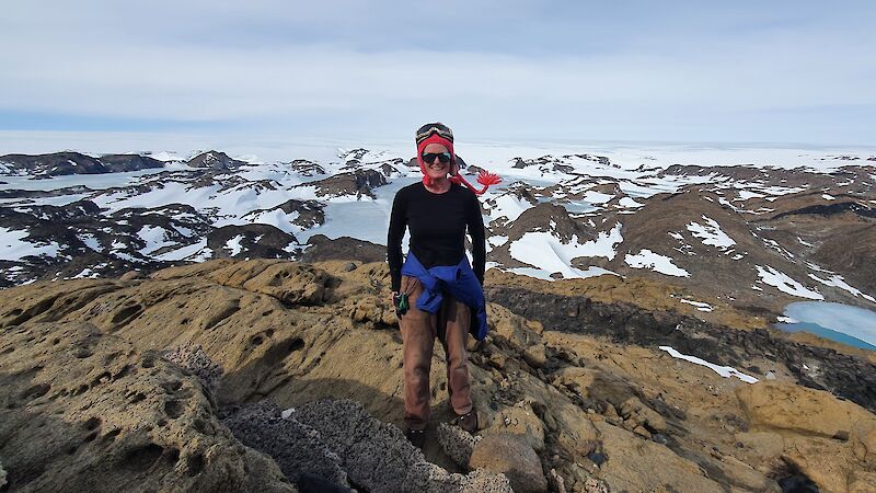 A woman with a red hat standing on a rocky outcrop with snow covered hills behind.