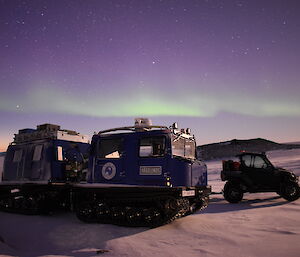 A blue Hägglund and Polaris vehicle in the snow.  The sky is lit up by a green aurora and orange sunset