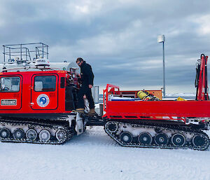 A red Hagglund vehicle and trailer in the snow. A man is perched at the back of the cab refuelling.