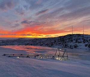 A snowy landscape with a ramp half covered in snow leading to a small lake.  The sky is orange and red as the sun rises behind the hill.