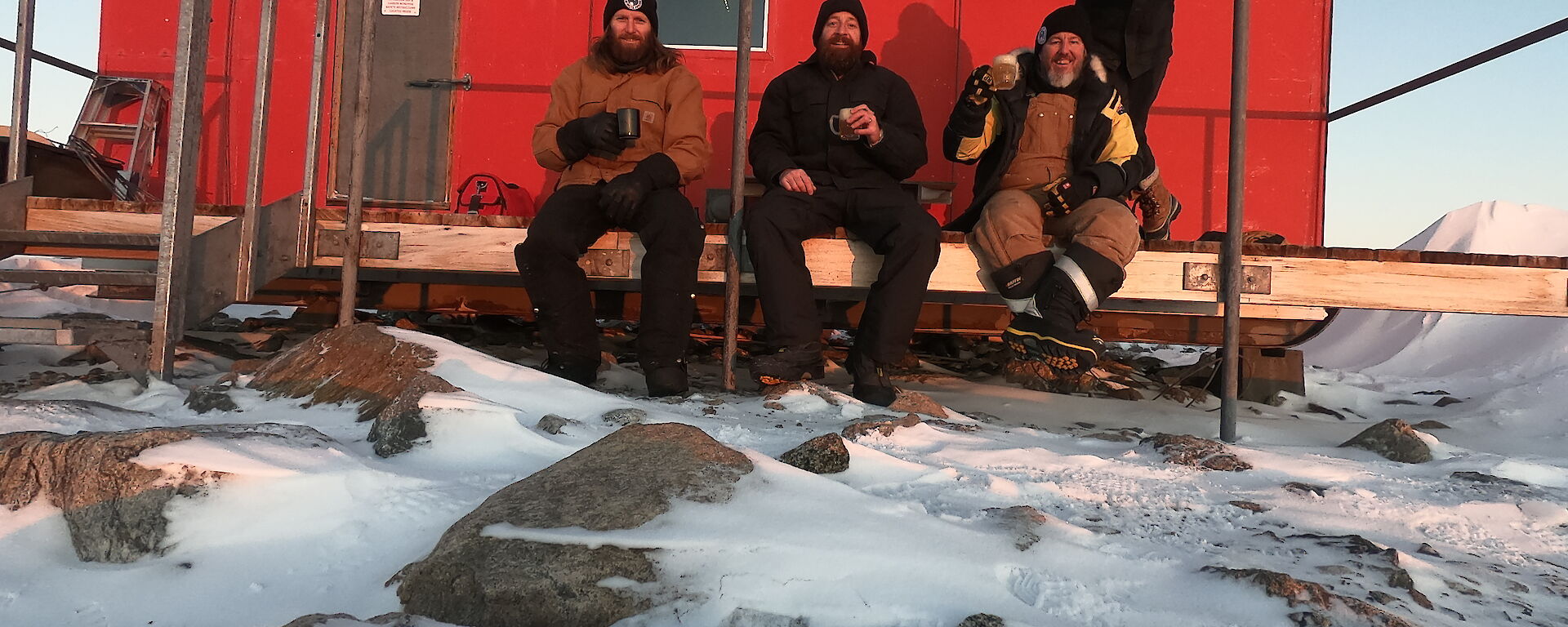 A red hut in the snow with three men sitting on a small veranda and another standing behind, all smiling to camera.