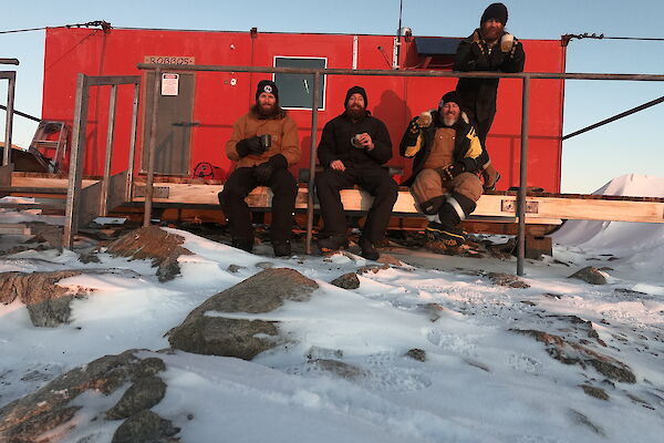 A red hut in the snow with three men sitting on a small veranda and another standing behind, all smiling to camera.