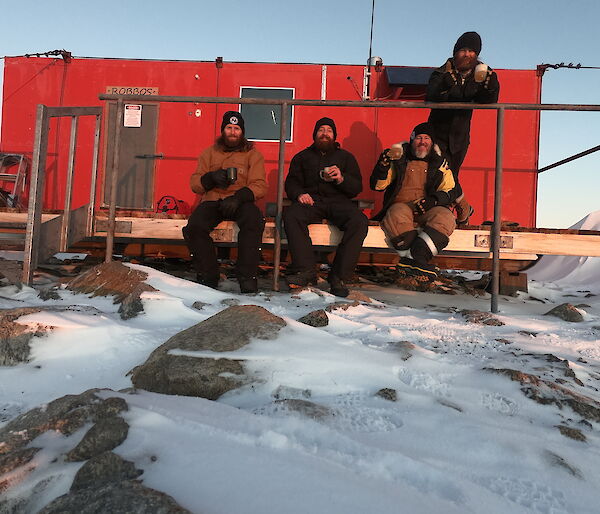 A red hut in the snow with three men sitting on a small veranda and another standing behind, all smiling to camera.
