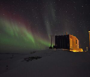 A green and purple aurora in the sky behind a shed, lit from the side by a lamp.