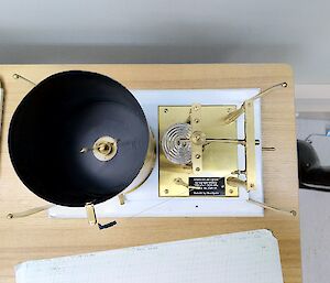 Looking down on to some weather observing equipment on a benchtop.  There is a cylinder on the left with some intricate measuring levers on the right.  Graph paper can be send at the bottom of the image.