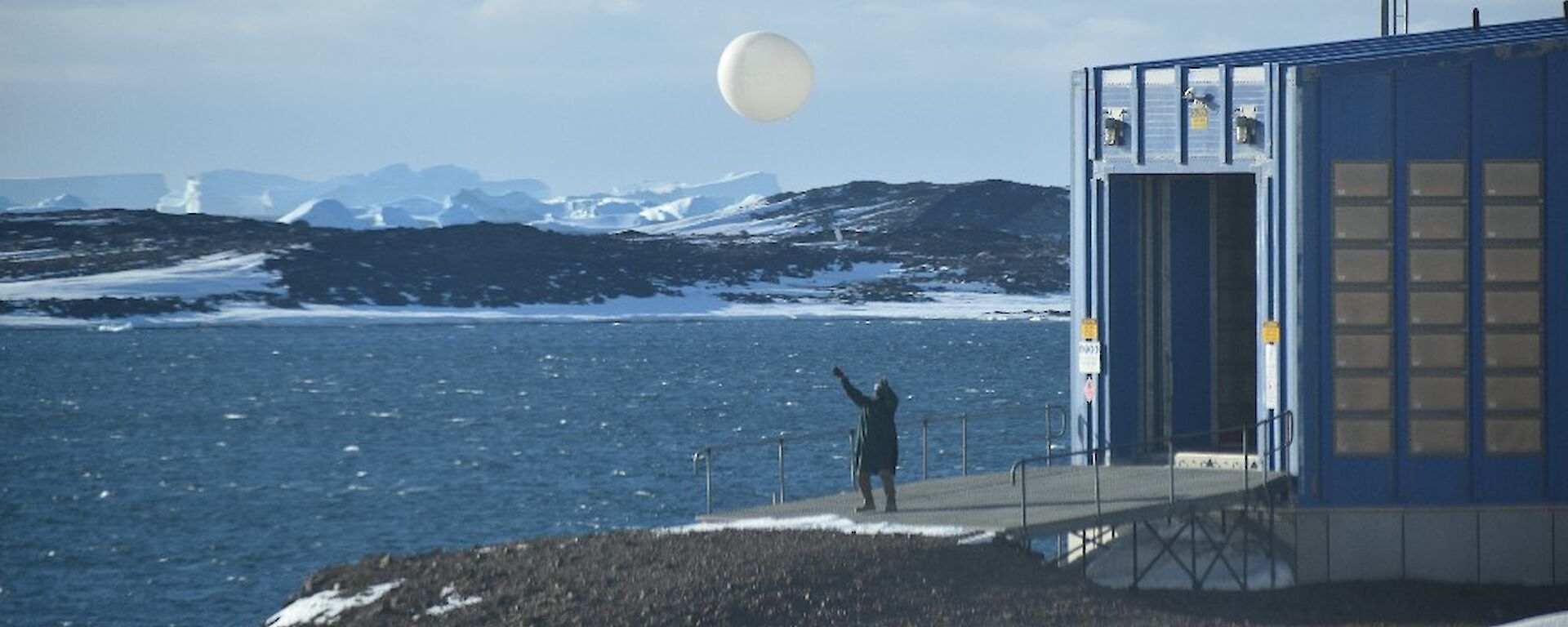 A man releasing the weather balloon just outside a shed at the edge of the water.  Ice capped mountains in the background.