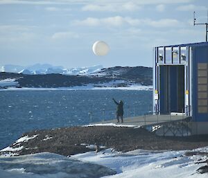 A man releasing the weather balloon just outside a shed at the edge of the water.  Ice capped mountains in the background.