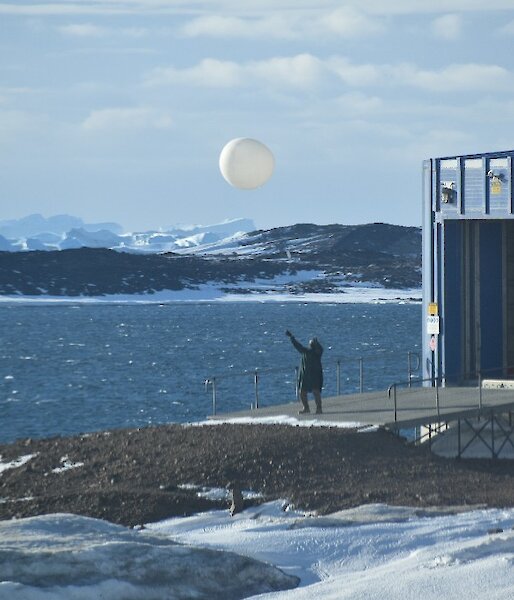 A man releasing the weather balloon just outside a shed at the edge of the water.  Ice capped mountains in the background.