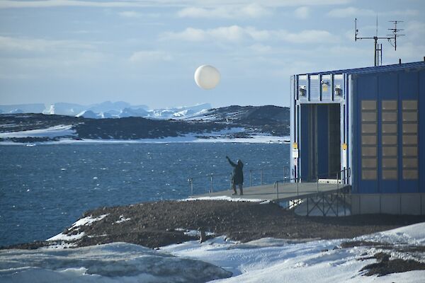 A man releasing the weather balloon just outside a shed at the edge of the water. Ice capped mountains in the background.