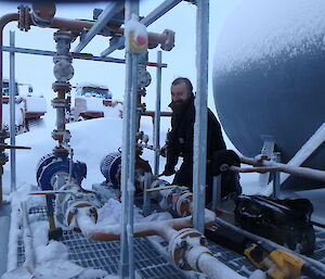 A man standing beside a series of pipes next to a large cylindrical tank.  The pipes and ground around him are covered in snow.