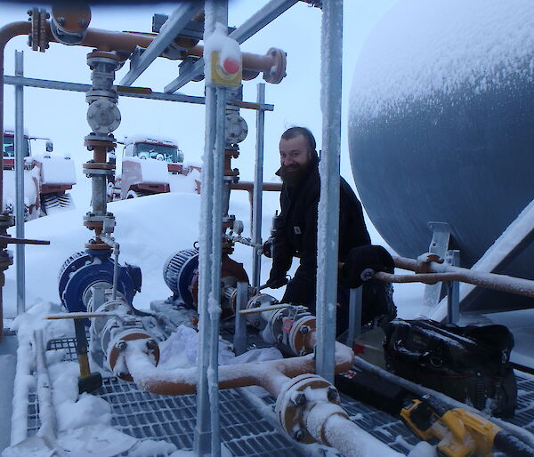 A man standing beside a series of pipes next to a large cylindrical tank.  The pipes and ground around him are covered in snow.