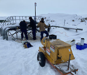 Three men in deep snow with a mobile generator fixing a burst pipe