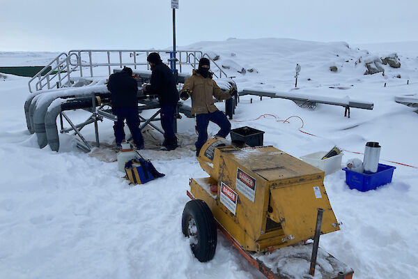 Three men in deep snow with a mobile generator fixing a burst pipe